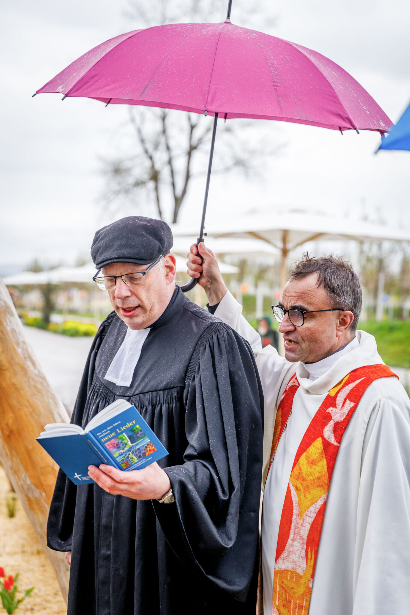 ein evangelischer und ein katholischer Geistlicher, beide singen, einer hält das Gesangbuch, einer hält den Regenschirm über beide