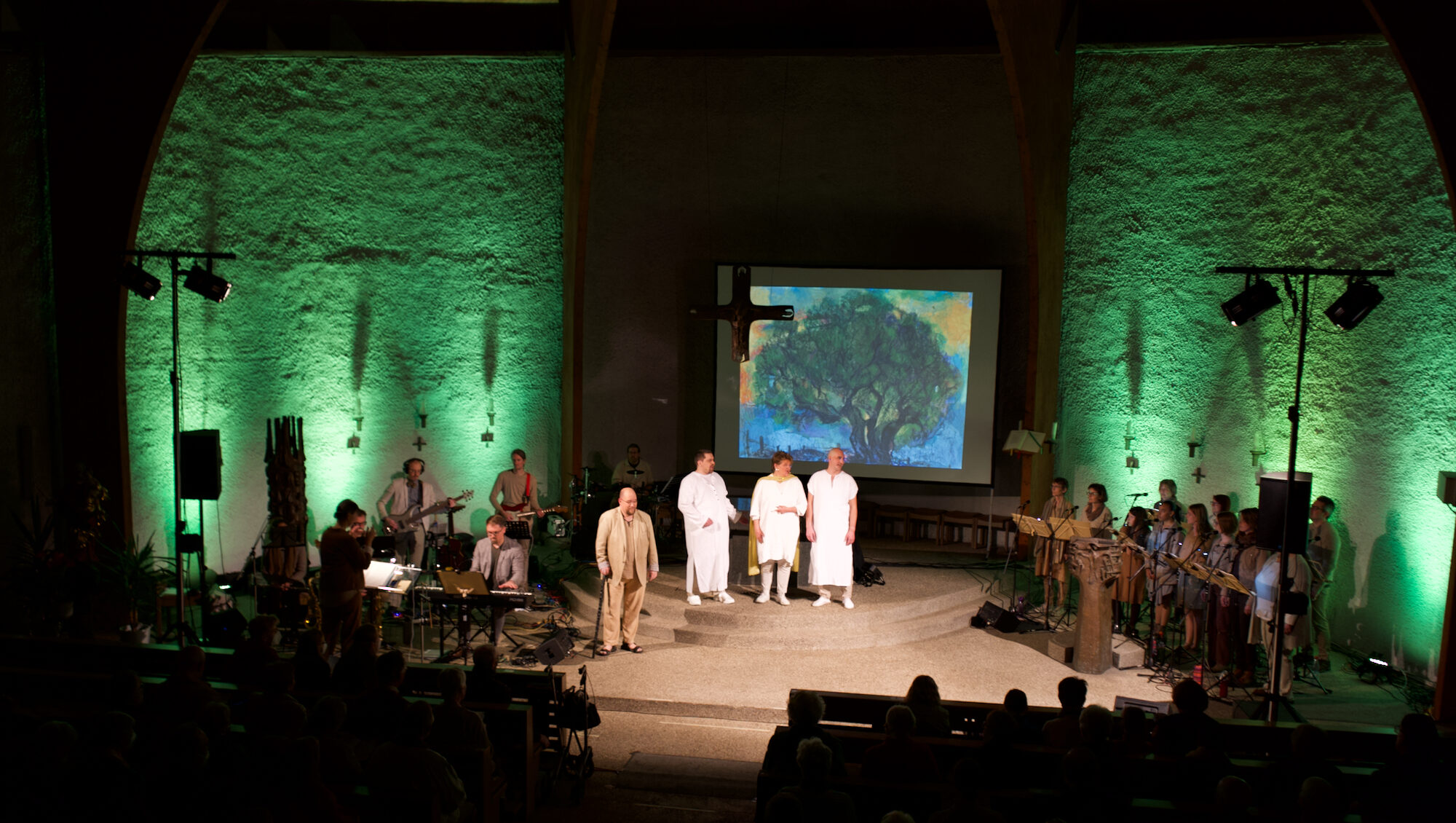 Kirchenraum grün illuminiert, Bild mit Baum im Hintergrund, davor 3 weiß gekleidete Männer, davor ein alter Mann, umrundet ist die Szene von Chor und Orchester