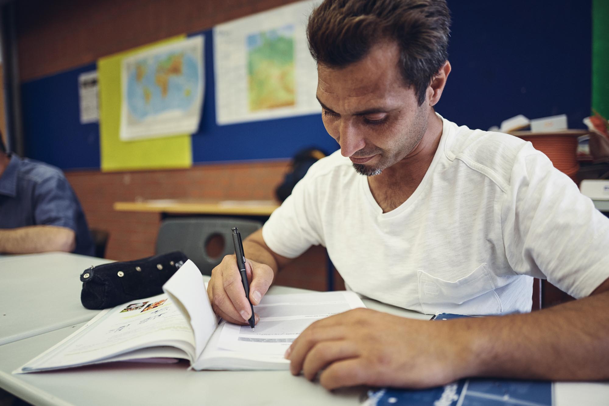 Ein Mann im weißen T-shirt sitzt an einem Stich in einem Klassenraum vor einem Buch und schreibt auf ein Arbeitsblatt