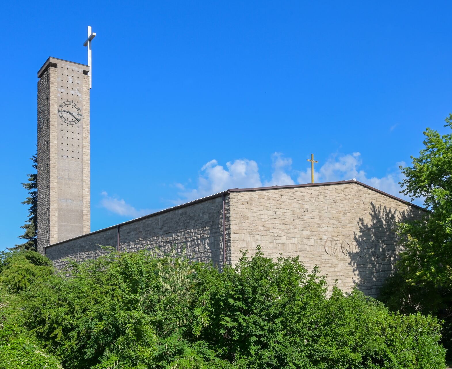 moderne Kirche von außen, grauer Stein, hoher, flacher Turm mit Kreuz und Uhr