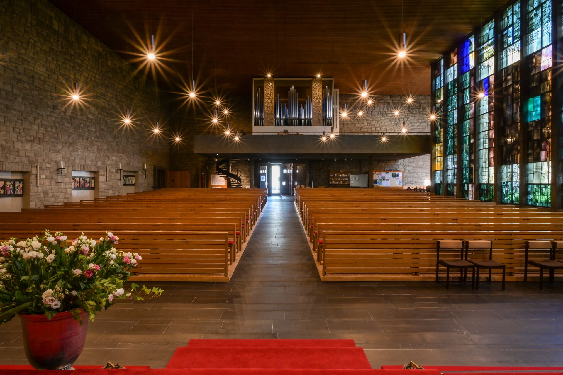Blick durch einen Kirchenraum auf eine Orgel, Lampen wie Sterne, bunte, moderne Glasfenster rechts im Bild