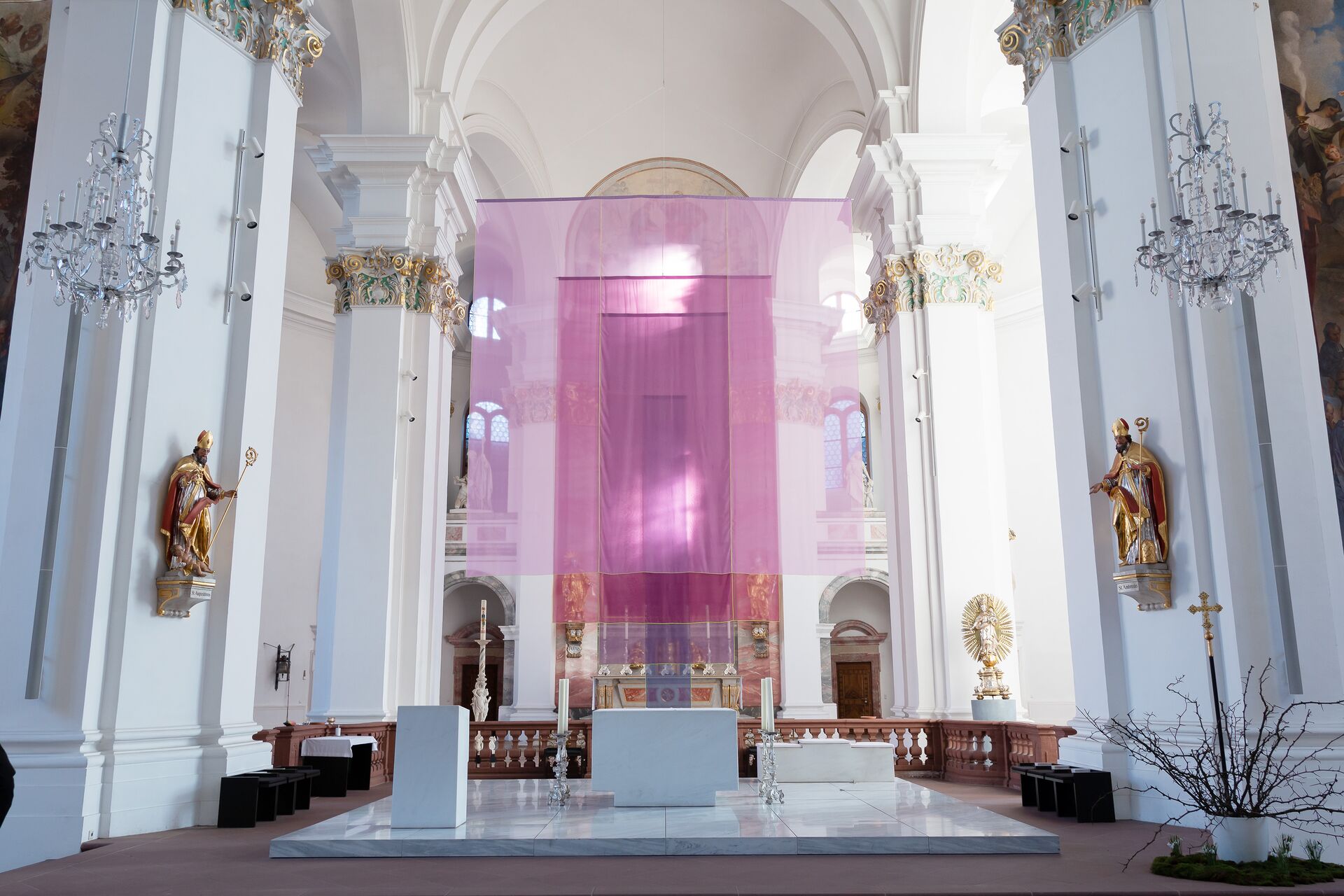 Blick in den Innenraum einer barocken, weißen Kirche, vorn im Altarraum verhüllt ein zartes, transparentes Tuch in violetten Farbtönen den Altar.
