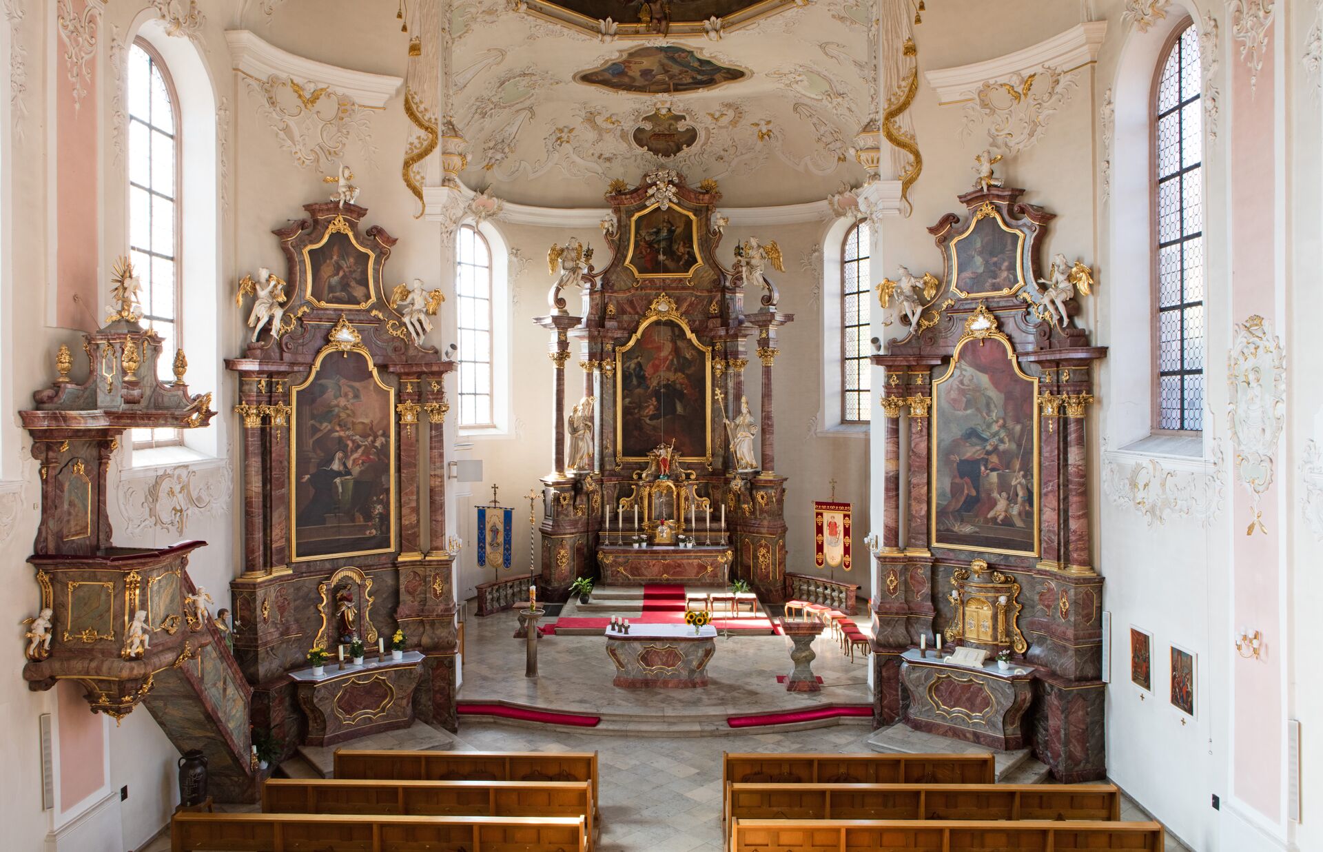 Blick in den Chorraum einer barocken Kirche, 3 Altäre, reich verzierte Stuckwände und Decken in weiß, rosa und gold