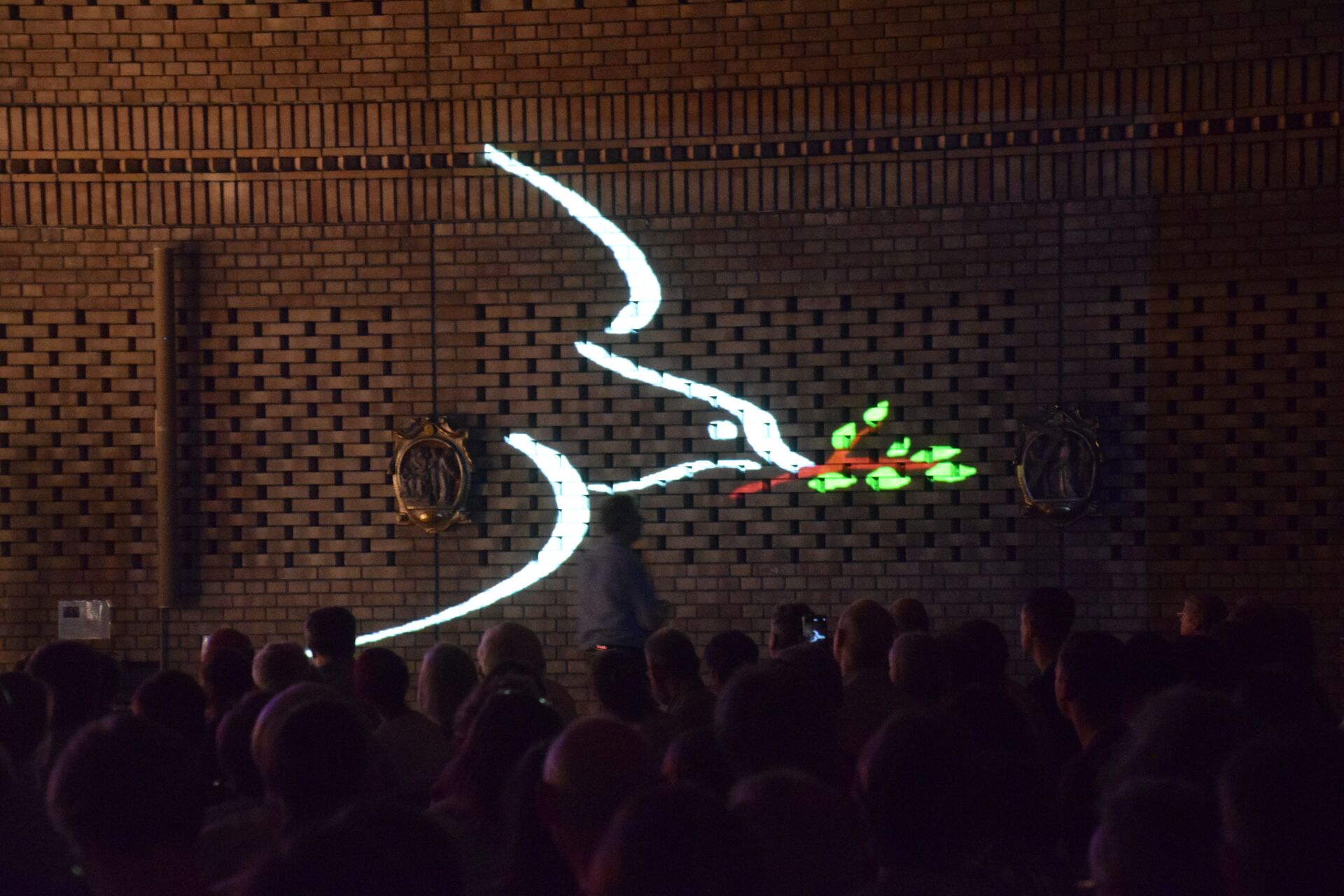 mit Licht an eine Kirchenwand aus gelben Backstein projizierte Friedenstaube, weiße Silhouette mit grünem Zweig mit Blättern