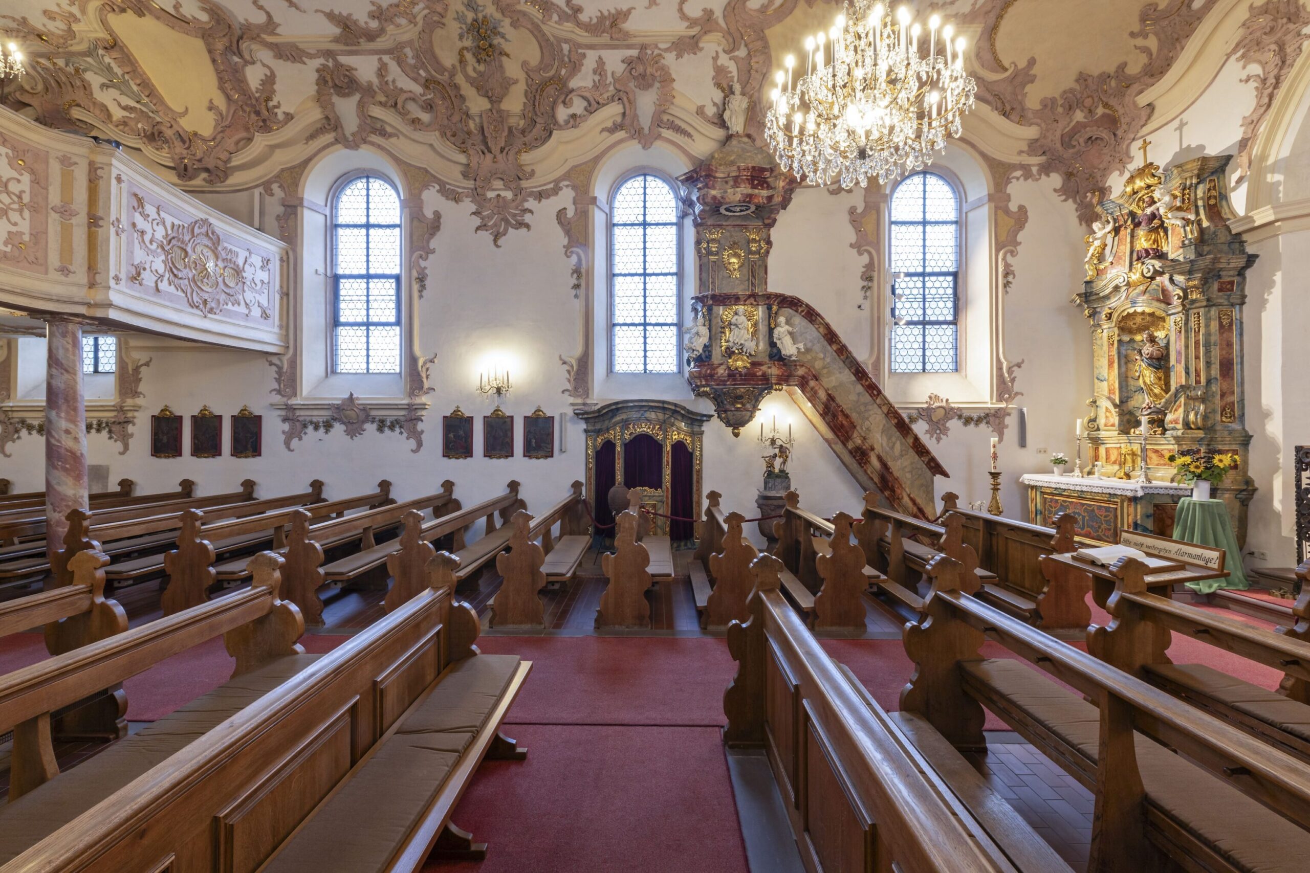 seitlicher Blick in eine Kirche mit Kirchenbänken und reicher Verzierung an den Wänden und Kronleuter