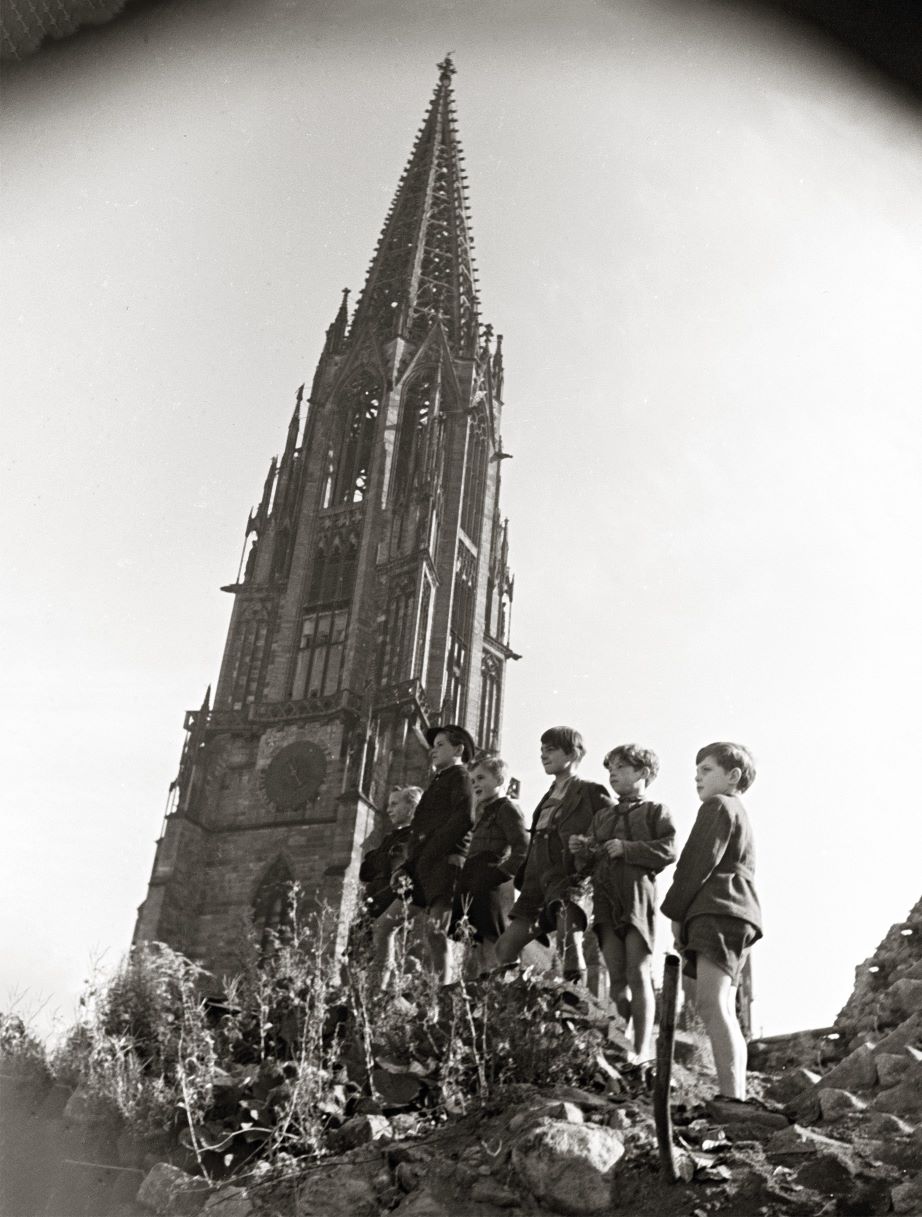 hoher Kirchturm, davor ein Schuttberg, darauf eine Gruppe von 6 Kindern still in einen Richtung schauend