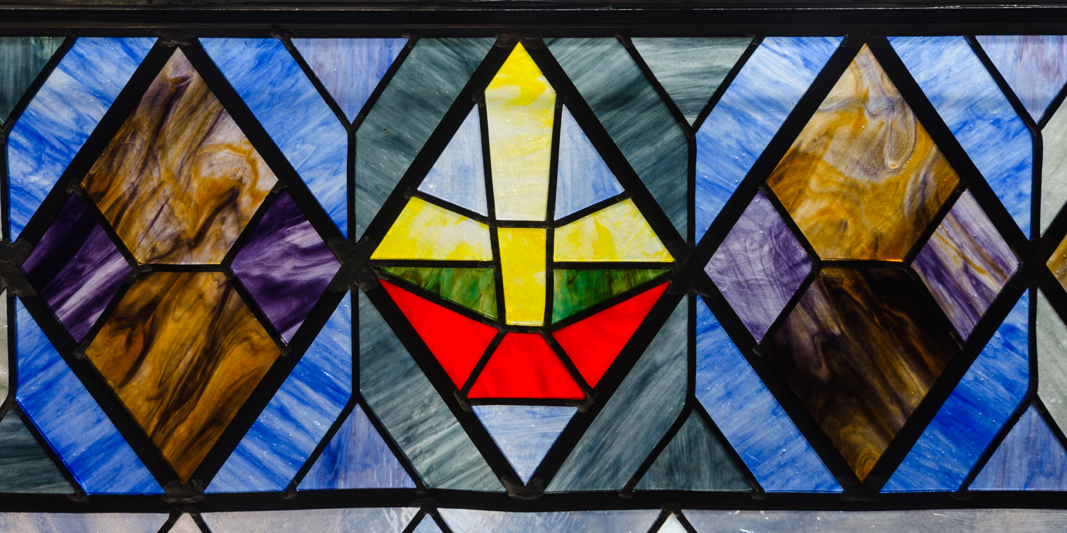 bunte Bleiglasfenster, leuchtende Blau, in der Mitte ein gelbes Kreuz oder Segel auf rotem Grund (Boot?)