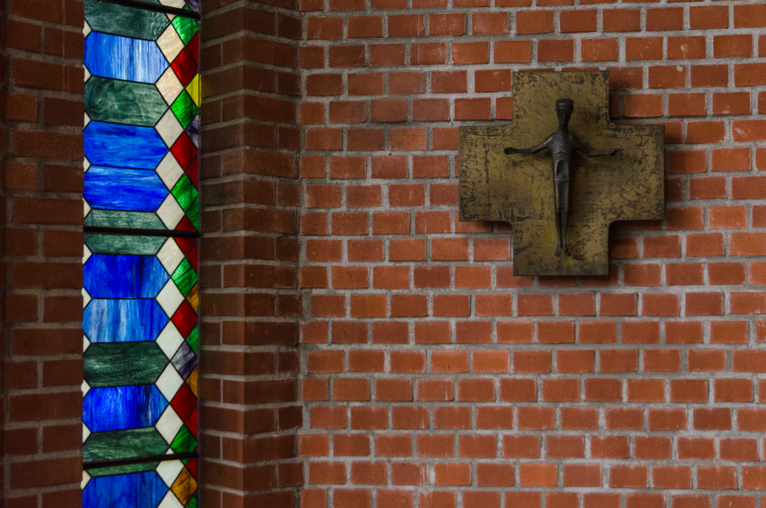 Ziegelwand mit Kreuz, links schmales Fenster aus Bleiglas in leuchten Blauen Ornamenten und weißen, roten, grünen Quadraten