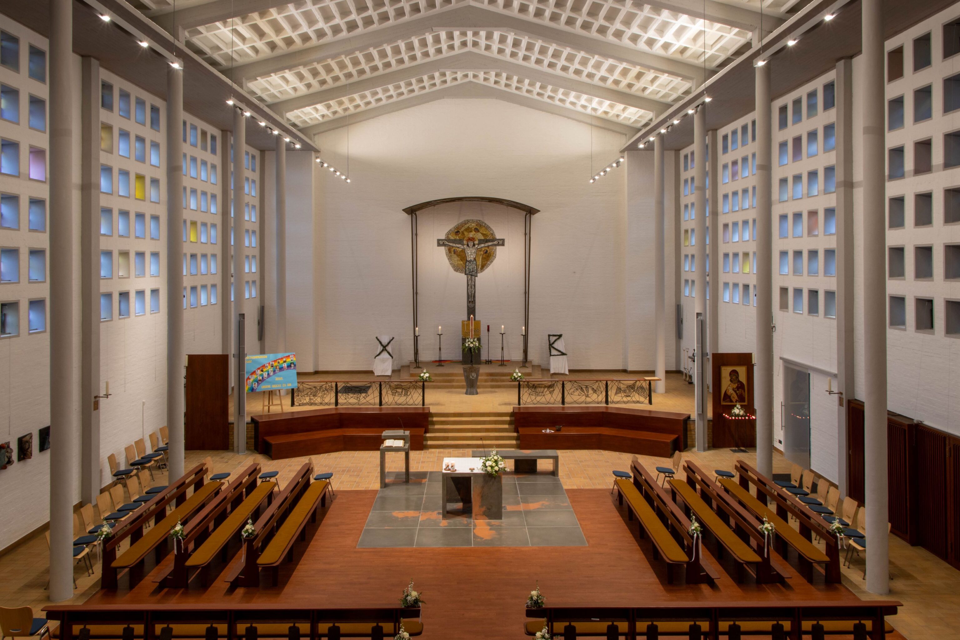Blick in eine große, moderne Kirche mit schmalen Säulen. Wie ein Halle. Der Altar steht in der MItte, die Bänke darum herum. Hinten an der Wand ein sehr großes Mosaikkreuz.