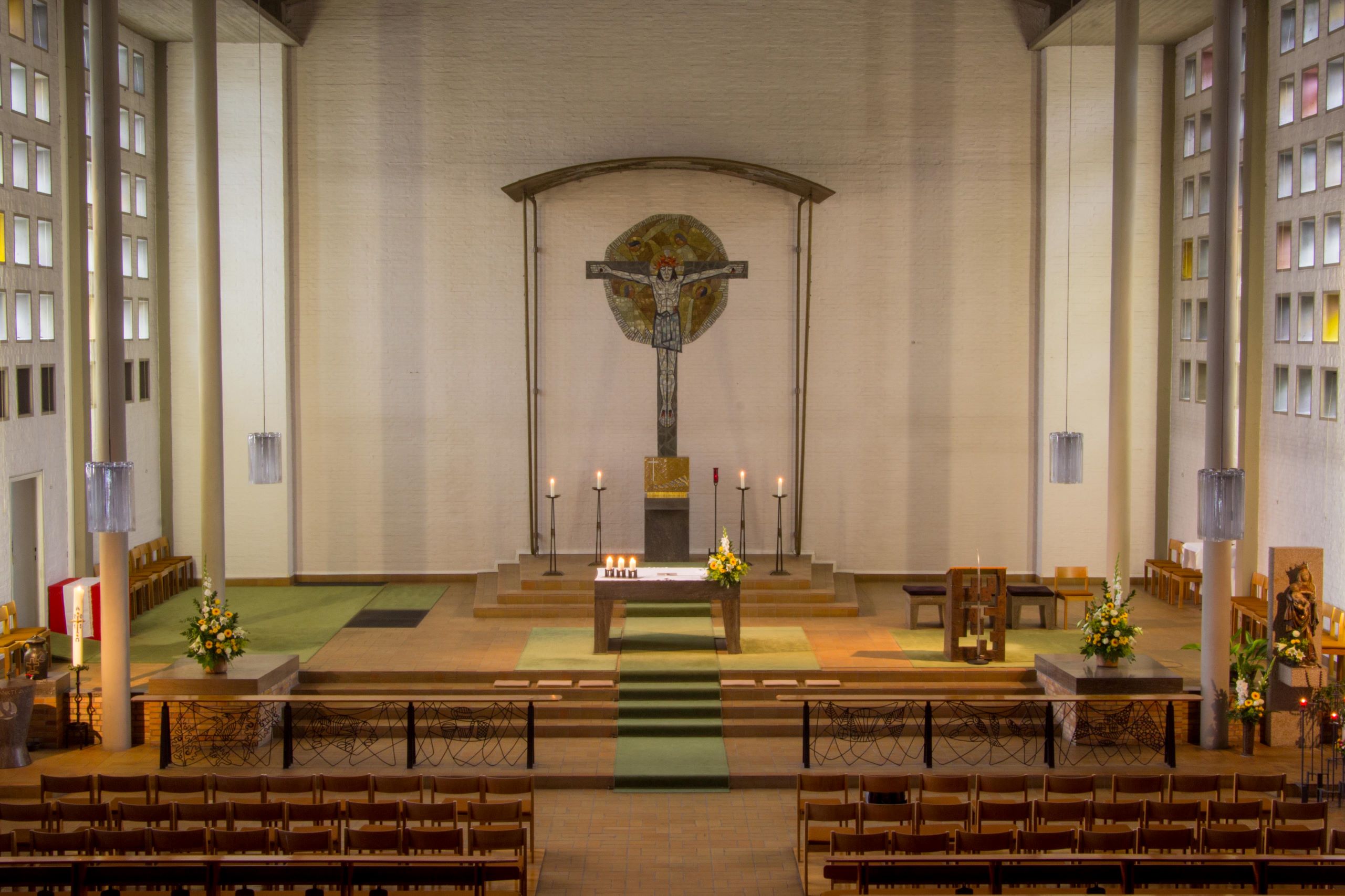 Großes Mosaikkreuz an der Wand im Chor, davor ein schlichter Altar, Bestuhlung gerade rechts und links davor im Kirchenschiff