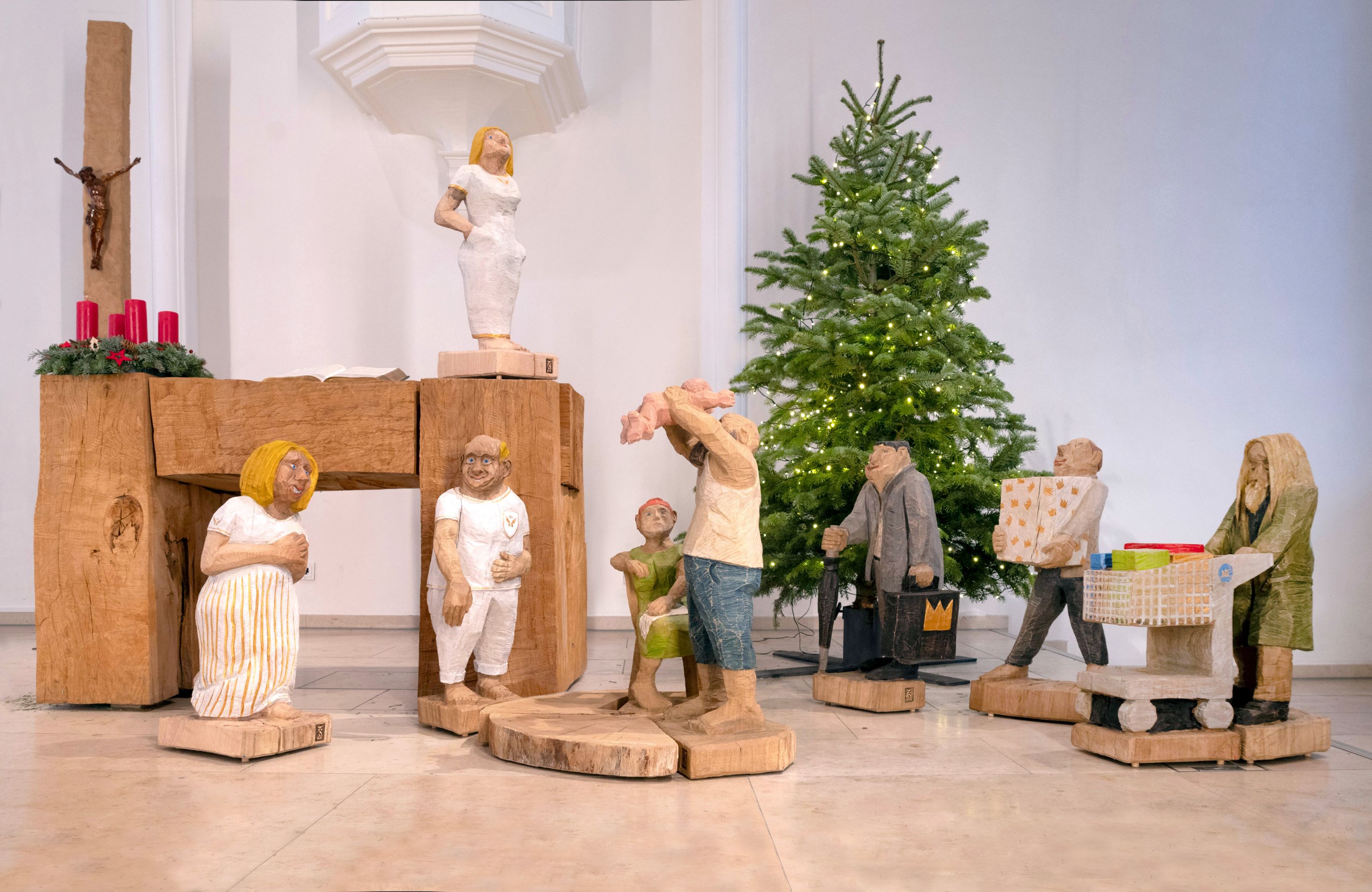 Krippenfiguren grob geschnitzt, in der heutigen Zeit mit einem Mann mit Einkaufswage, mit Paket und Einkaufstüte, Josef in Jeans und Unterhemd hält das nackte Kind hoch über seinen Kopf, Maria sitzt erschöpft auf einem Stuhl daneben,