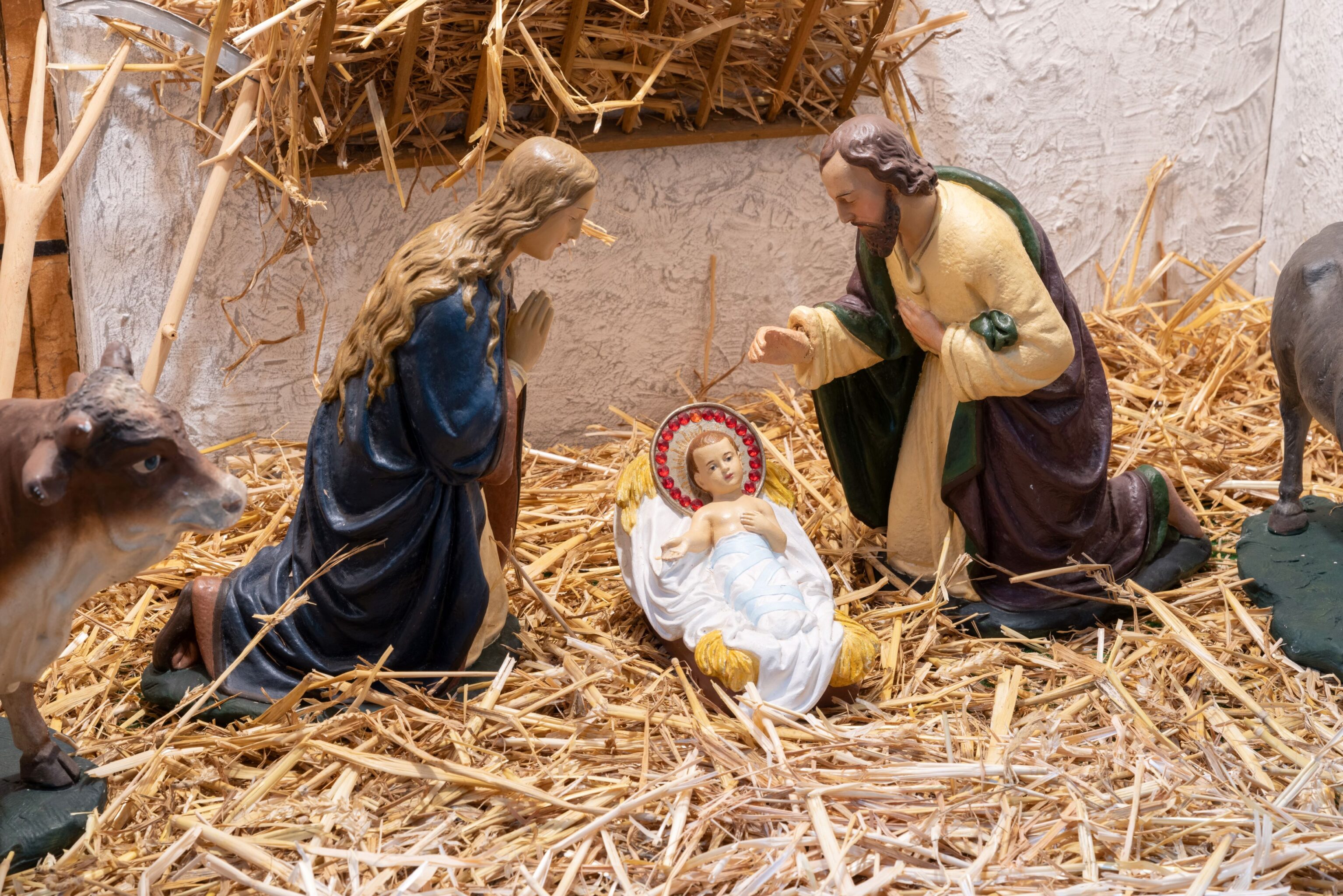 Maria und Josef knien neben dem Jesuskind. Dieses hat einen auffälligen Heiligenschein mit roten Perlen. Die Figuren stehen auf Stoh.