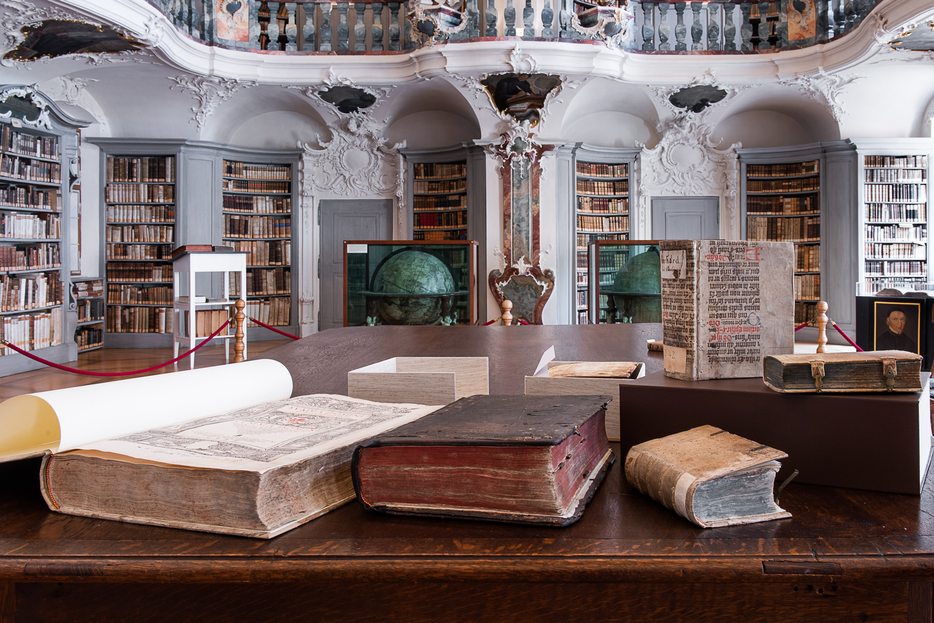 sehr alte, dicke Bücher auf dunklem Holztisch in einer hellen, barocken Bibliothek, hohe Bücherregale
