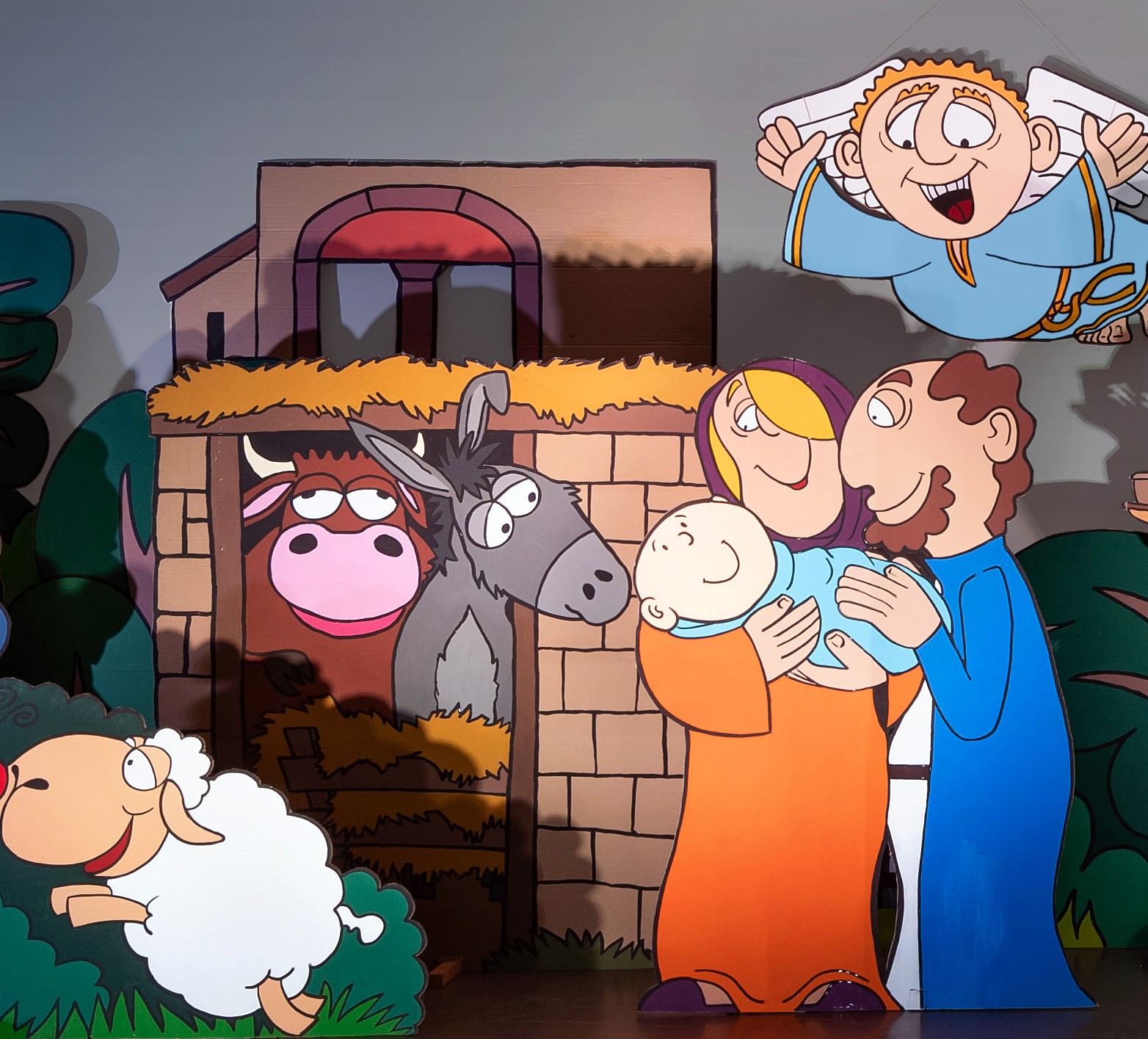 Krippe als Cartoon lustig und bunt, Maria und Josef mit Kind, darüber ein Engel, Ochs und Esel schauen aus dem Stall heraus, ein weißes Schaf springt davon