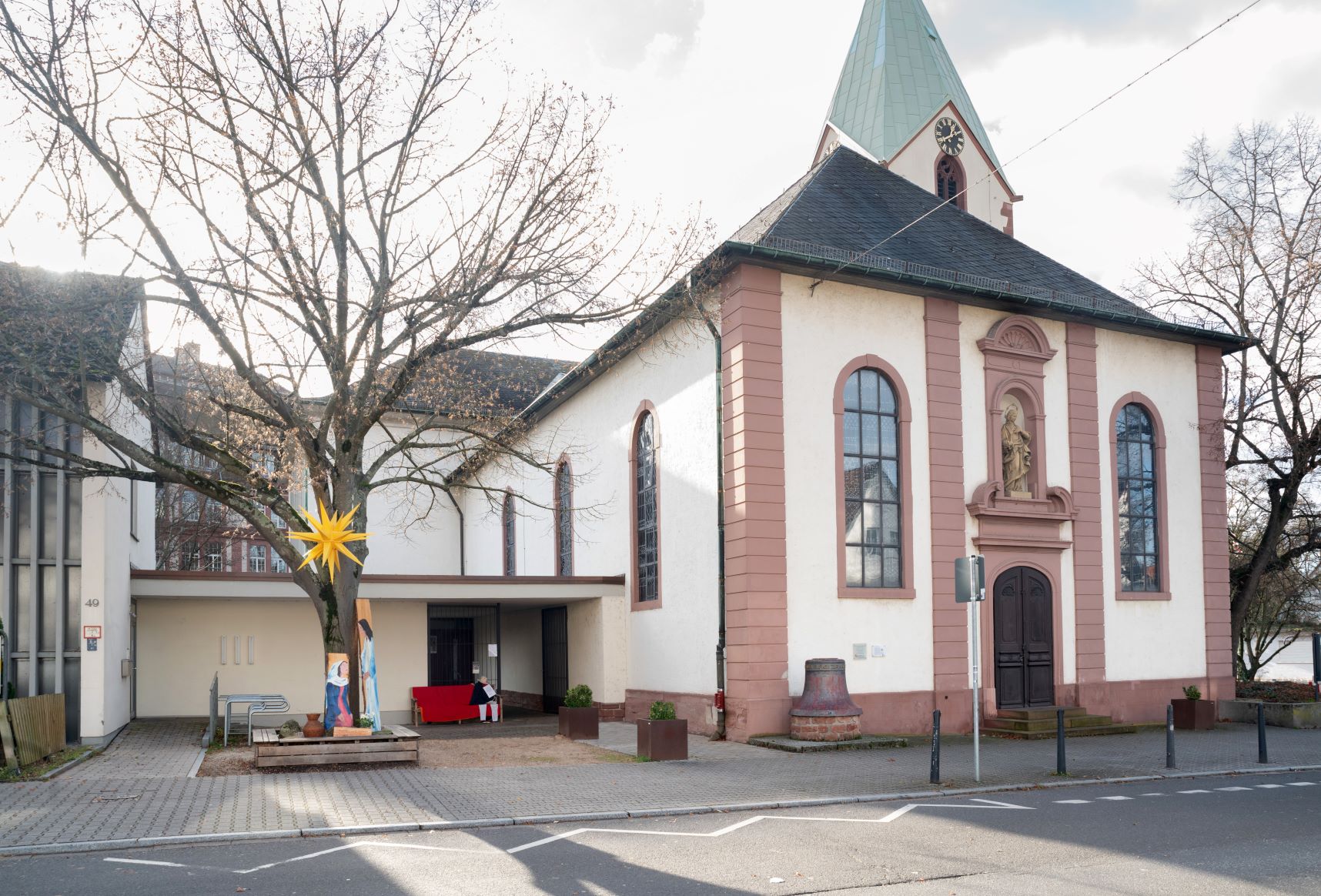 Kirche von aussen, weiß und Sandstein, links davon ein Platz mit Baum im Winter, daran ein gelber Stern, barrierefreier Zugang in die Kirche