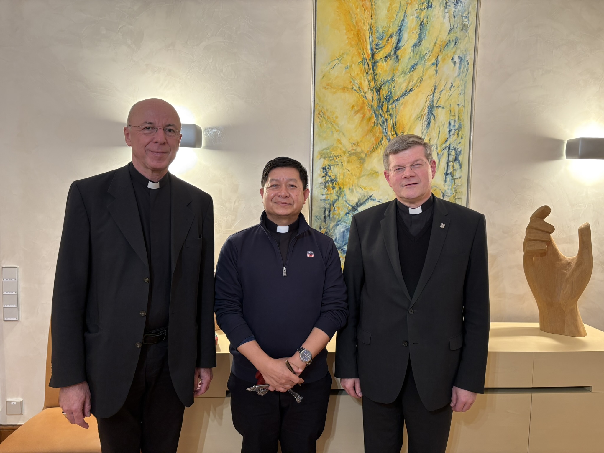 Drei Priester nebeneinander in Schwarz gekleidet, im Hintergrund ein modernes Gemälde in gelb,blau, daneben eine geschnitze, offene Hand