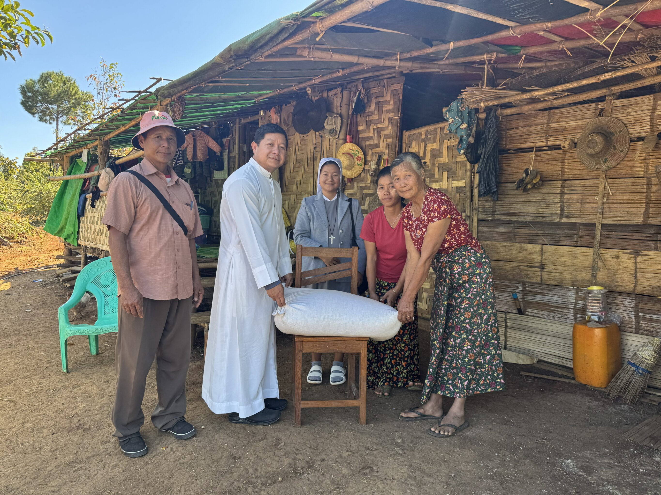 Fünf Personen vor einem einfachen Haus aus Holz und Bambu, In der Mitte ein Reissack, ein Priester überreicht diesen an zwei Frauen