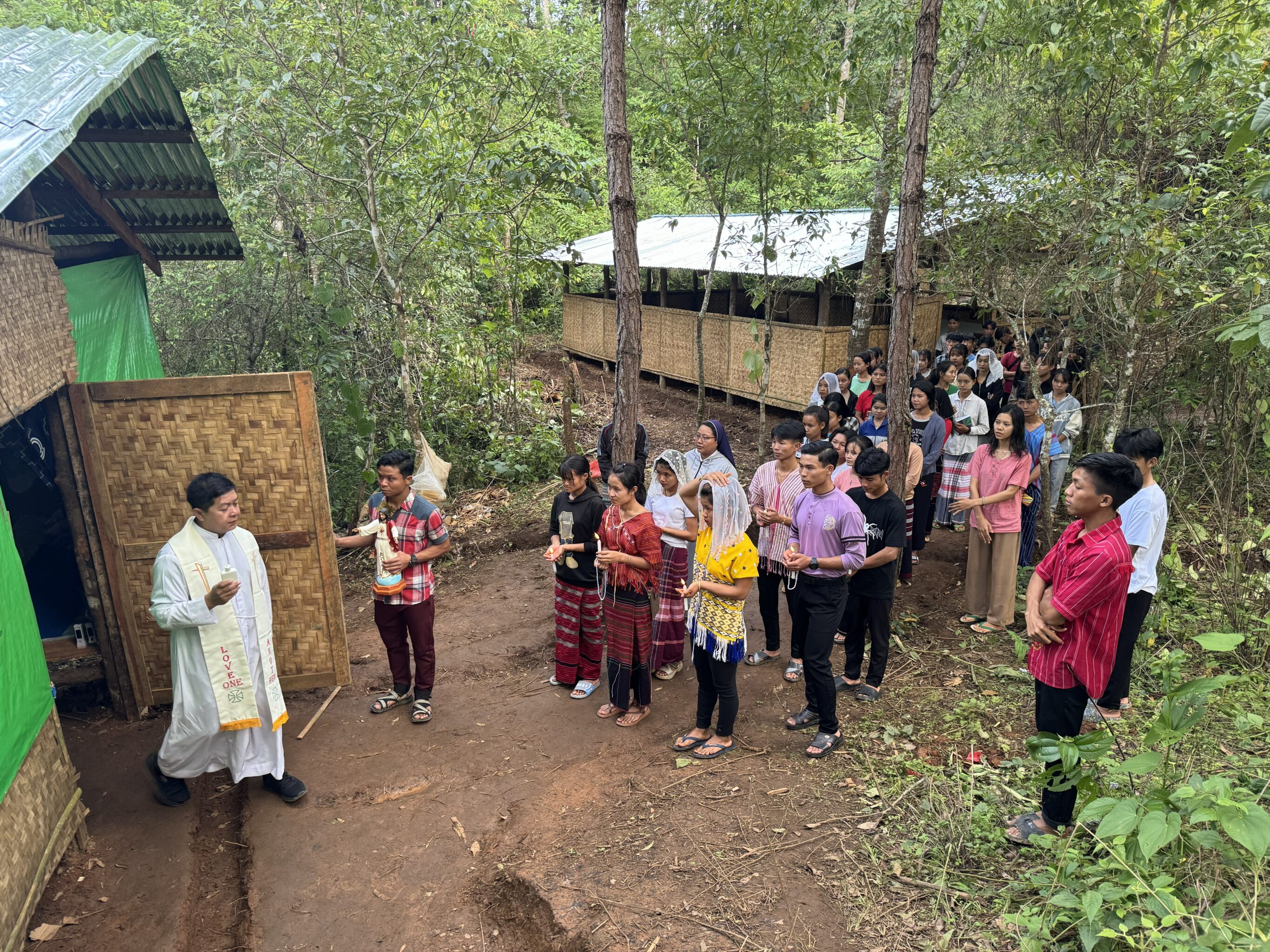 Gottesdienst unter freiem Himmel im Wald, die Meschen stehen, der Priester kommt einer Hütte dazu