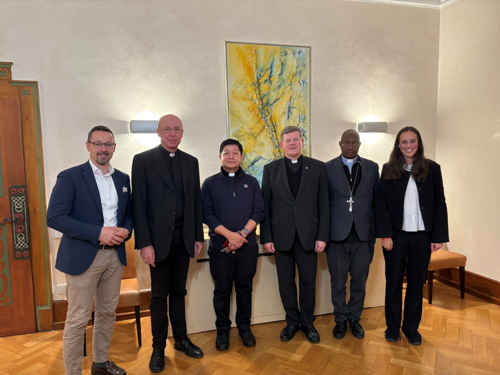 Fünf Männer, davon vier Priester, und eine Frau nebeneinander stehend in die Kamera schauend, im Hintergrund ein modernes Gemälde in gelb,blau,