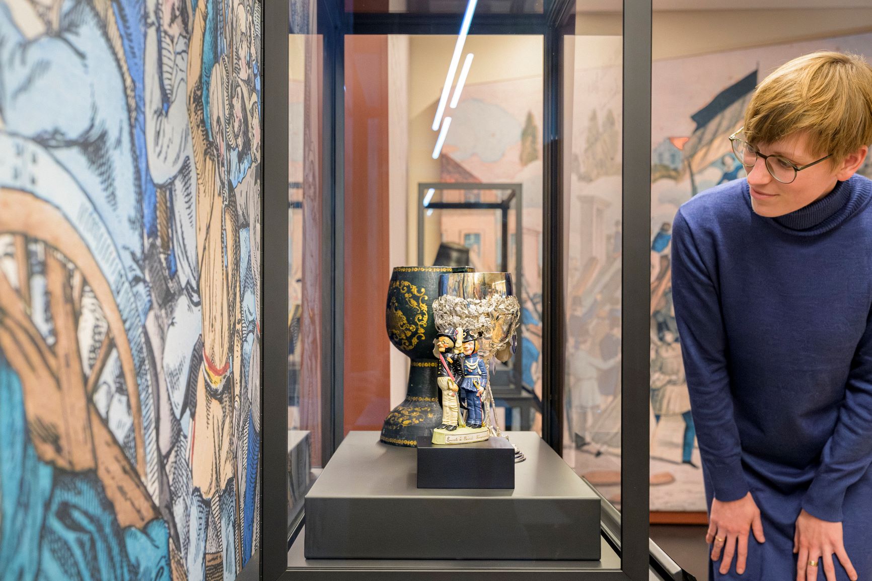 eine Frau in blauem Pullover beugt sich vor eine Vitrine, in der ein großer Kelch und kleine Figuren stehen.
