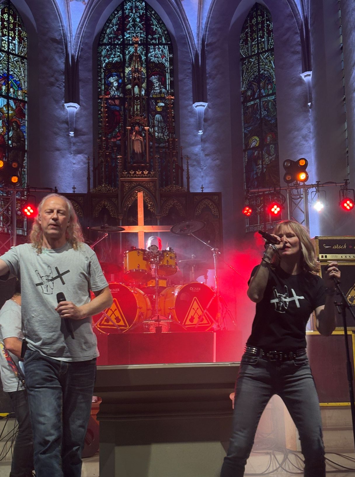Ein Sänger und eine Sängerin, im Hintergrund ein Schlagzeug, dahinter ein Kreuz, Nebel im roten Licht, im Chorraum einer Kirche, hinten bunte gotische Kirchenfenster, auf den T-shirts der Sänger die Hand mit Metal signe und Kreuz
