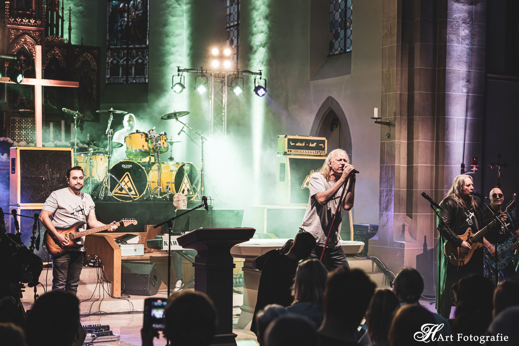 Musiker mit Schlagzeug, Bass, Verstärkern im Chorraum einer Kirche, Nebel und grünes Licht, im Hintergrund ein großes Kreuz und ein Altar, vorne Menschen in der Kirche