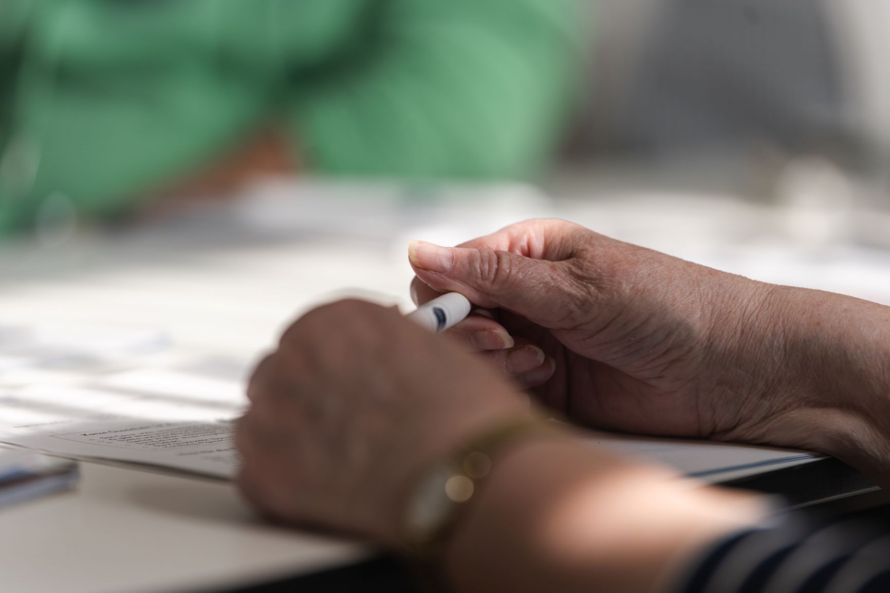 zwei Hände liegen auf einem Tisch auf Papieren, halten einen Kugelschreiber zwischen sich, gegenüber sitzt unscharf eine Person