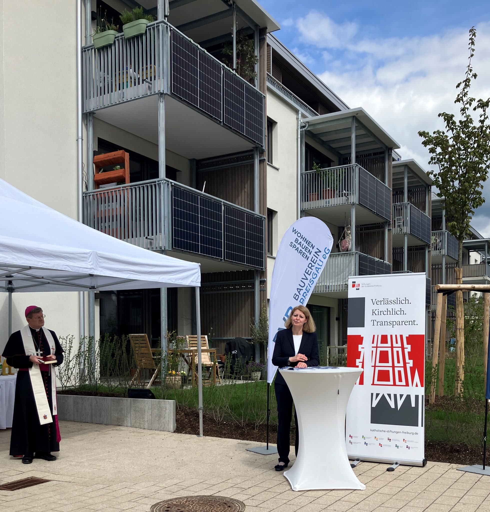 moderne 3-stöckige Häuser im Hintergrund, vorne unter einem weißen Pavillon Erzbischof Burger mit Stola, an eimem Stehtisch spricht Dana Mebus, dahinter Rollup mit Logo rot-grau-weiß
