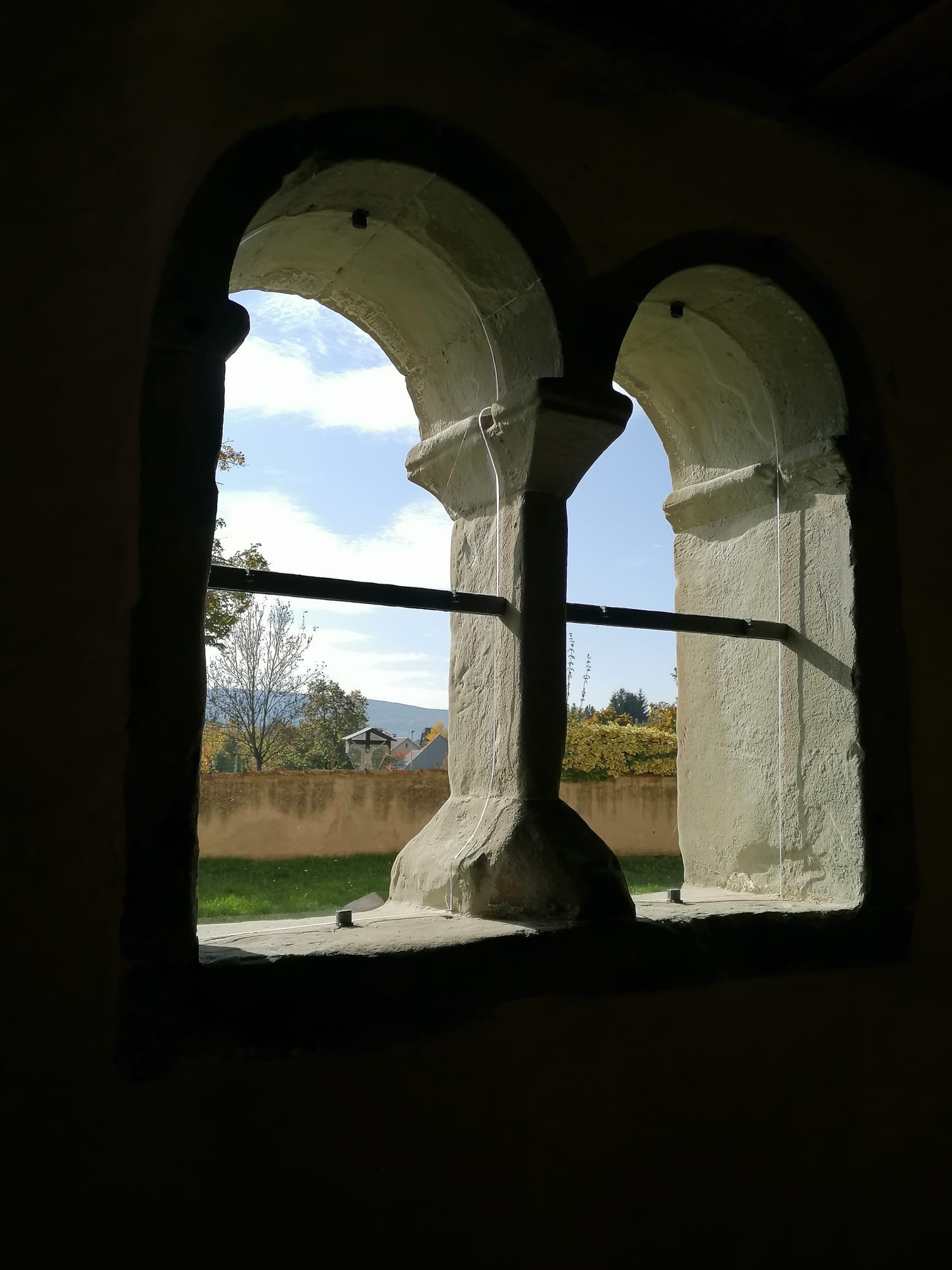Blick durch zwei leere Fensterbögen nach draußen auf eine Rasenfläche, Mauer und dahinter Bäume, ein Kreuz, Hügel