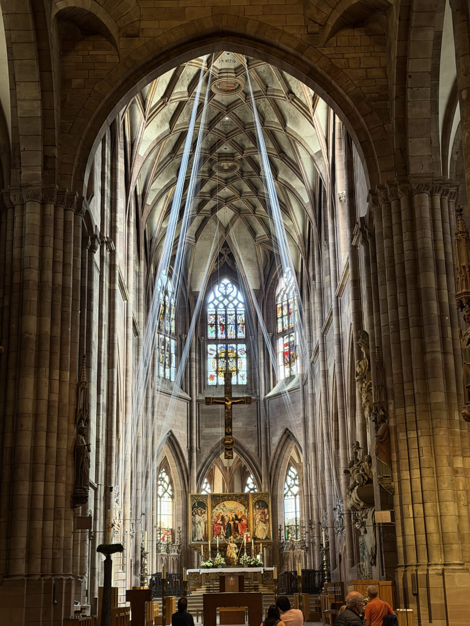 Blick in den Chorraum einer gotischen Kirche, von der Decke bis fast nach unten sind weiße Fäden gespannt, die wie Lichtstrahlen aussehen, hinten ein Altar, in der Mitte hängt ein großes Kreuz