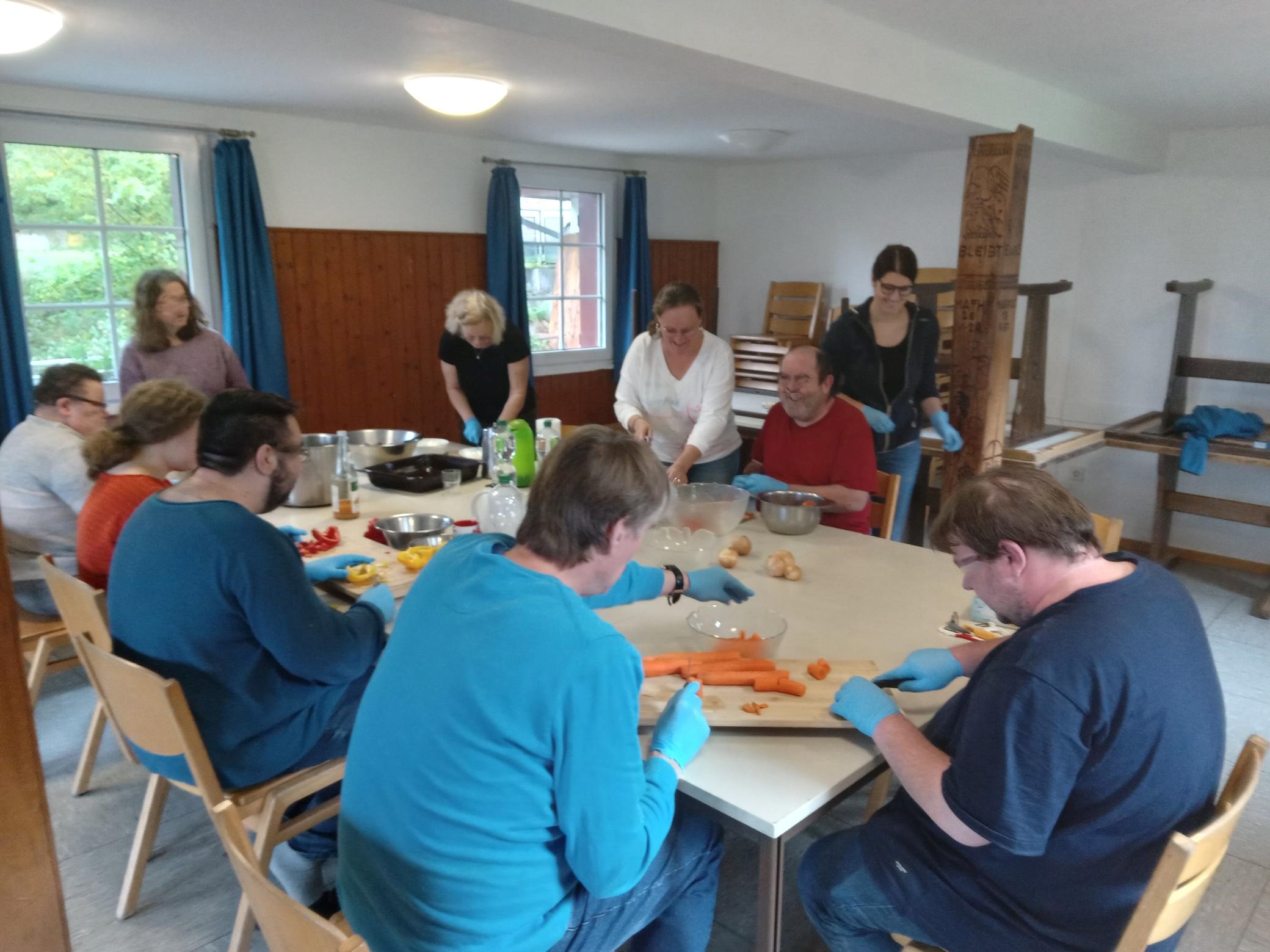 Zehn Menschen sitzen und stehen fröhlich um einen Tisch und schneiden Gemüse klein.