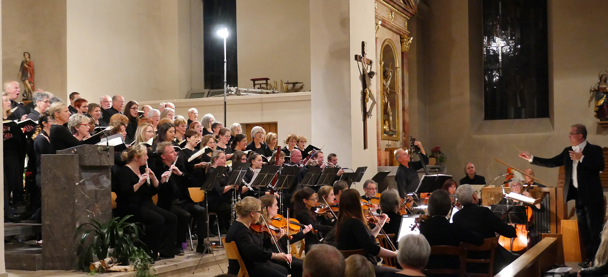 Chor und Orchester in einer Kirche, alle festlich schwarz gekleidet, auf der rechten Seite steht der Dirigent