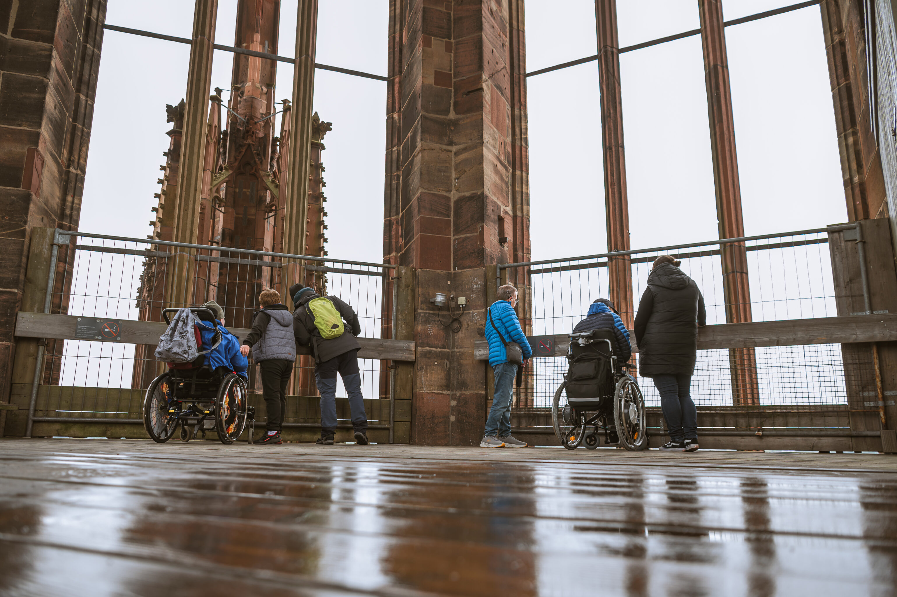 sechs Personen stehen auf der Plattforum des Münsterturms und schauen in die Ferne. 2 Personen sitzen im Rollstuhl. Der Boden ist nass.