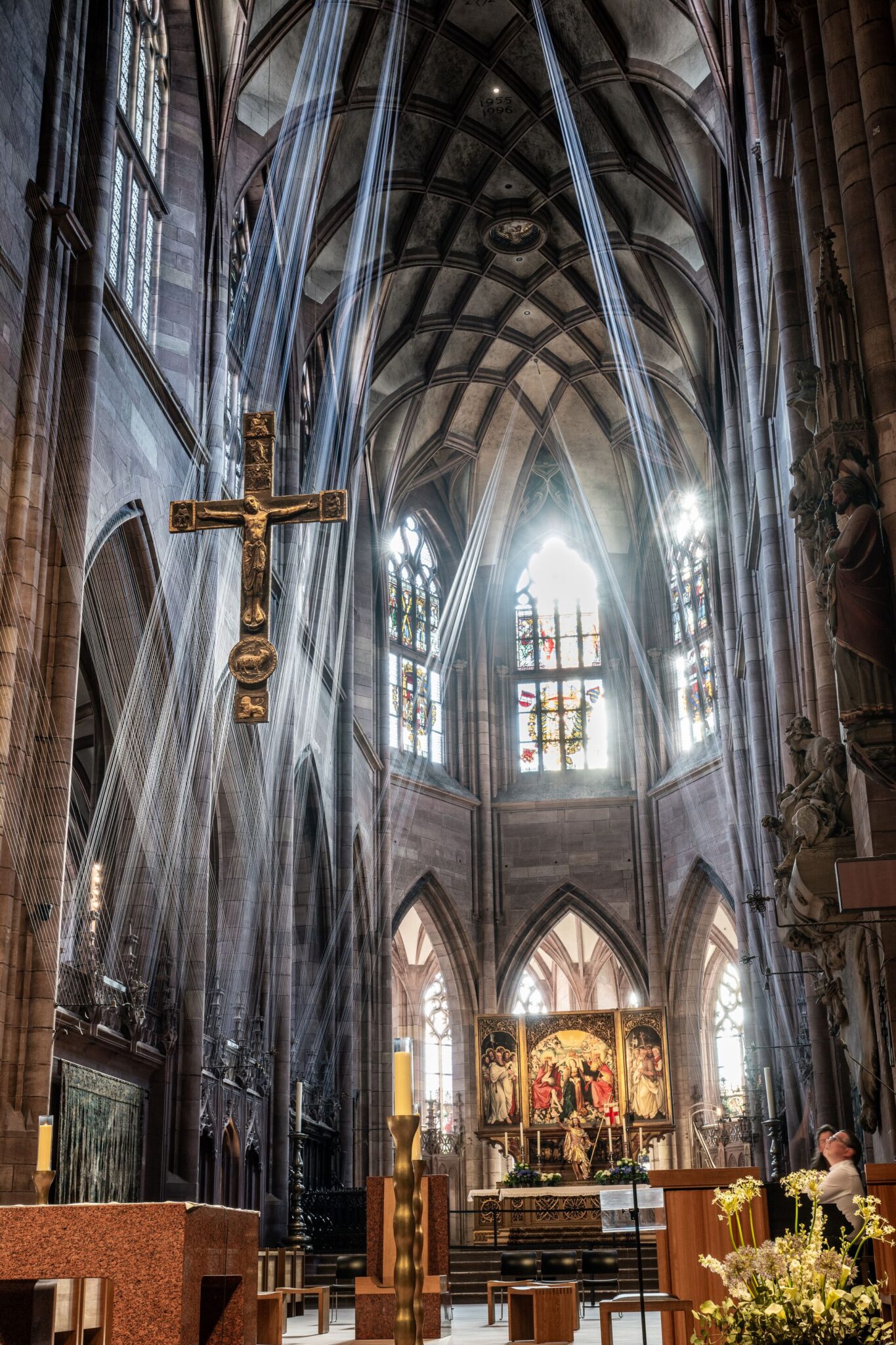 Blick in den Chorraum, Lichtdurchflutet, vorne hängt ein Kreuz von der hohen, gotischen Decke, durch den Raum sind, wie Lichtstrahlen, Fäden gespannt