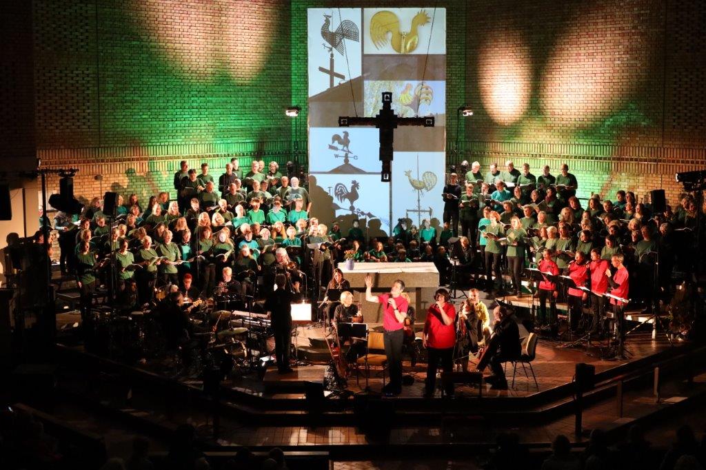 großer Chor hinten in grünen Shirt, vorne Solisten in roten Shirts, mit Orchester gruppiert im Altarraum einer Backsteinkirche, hinten eine Leinwand mit verschiedenen Motiven von Wetterhähnen, vorne hängt ein großes Kreuz