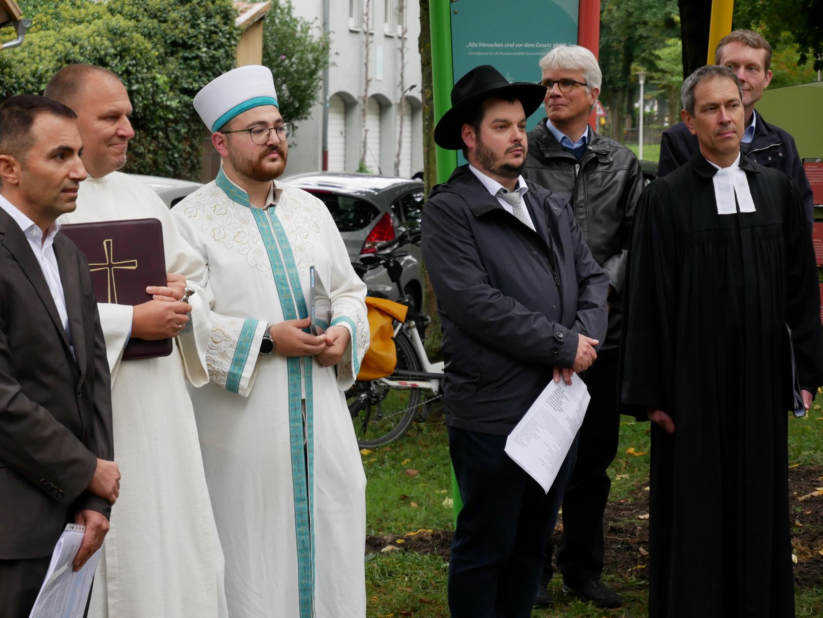 sieben Männer stehen draußen, zwei im Hintergrund. Vorne Mann in dunklem Anzug, ein katholischer Priester mit großem Buch mit Kreuz und ein muslimischer Geistlicher, beide in weißem Gewand, ein Rabbiner mit großem Hut und ein evangelischer Pfarrer in schwarzem Talar.